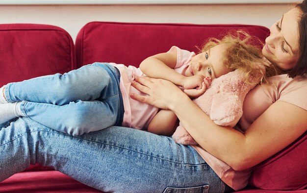 Foto liggend op rode bank jonge moeder met haar dochtertje in vrijetijdskleding samen binnenshuis thuis