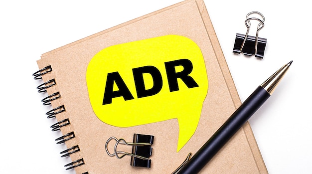 На светлом фоне коричневый блокнот, черная ручка, скрепки и желтая карточка с текстом ADR Alternative Dispute Resolution Business concept