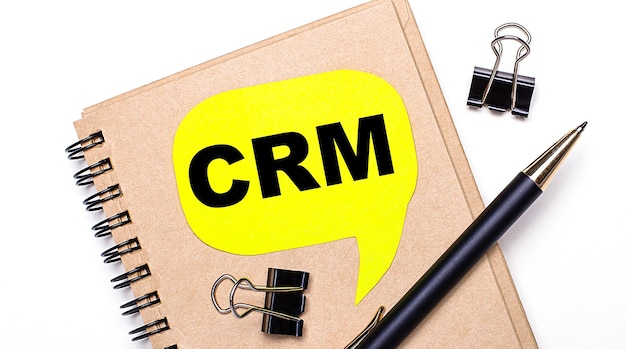 On a light background a brown notebook a black pen and paper clips and a yellow card with the text CRM Customer Relationship Management