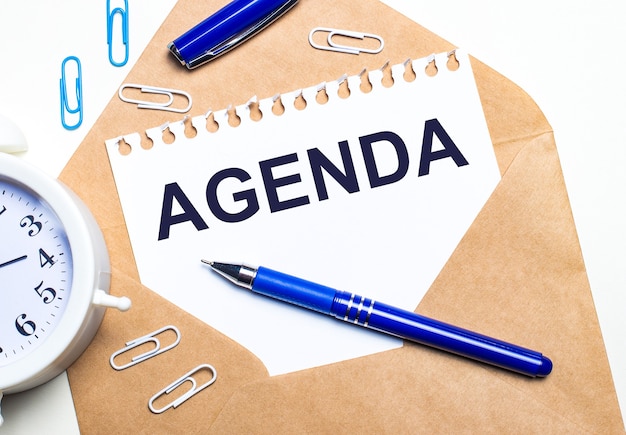 On a light background, a craft envelope, an alarm clock, paper clips, a blue pen and a sheet of paper with the text AGENDA.