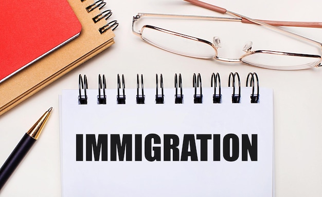 On a light background glasses in gold frames a pen brown and red notepads and a white notebook with the text IMMIGRATION Business concept