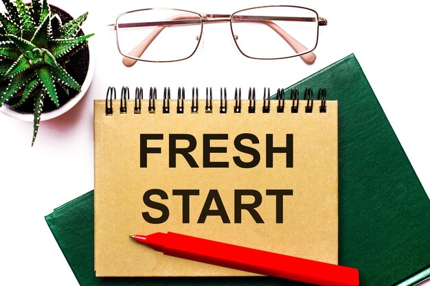 On a light background goldframed glasses a flower in a pot a green notebook a red pen and a brown notebook with the text FRESH START Business concept