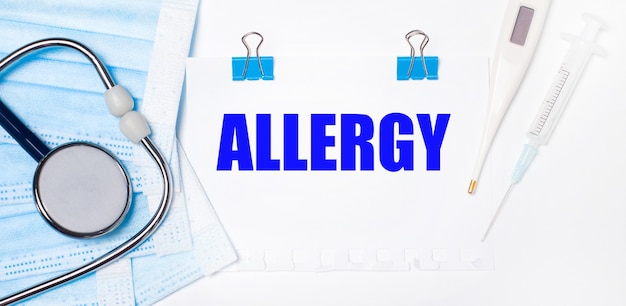 On a light background lie a stethoscope, an electronic thermometer, a syringe, a face mask and a sheet of paper with the text ALLERGY. Medical concept