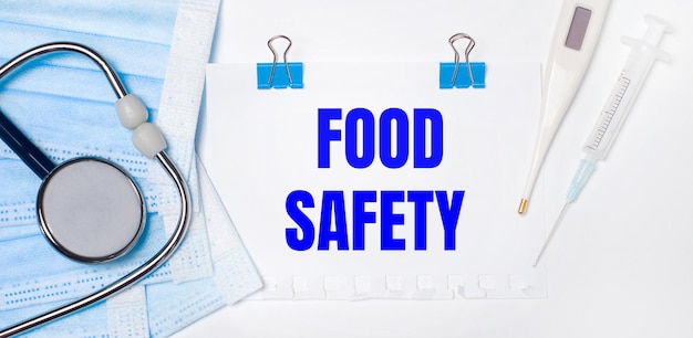 On a light background lie a stethoscope, an electronic thermometer, a syringe, a face mask and a sheet of paper with the text FOOD SAFETY. Medical concept