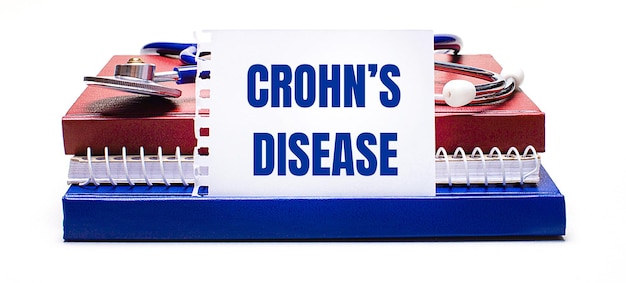 明るい背景のメモ帳に聴診器と白いカードに「CROHN'SDISEASEMedicalconcept」というテキストが表示されます