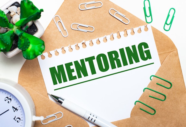 On a light background, an open envelope, a white alarm clock, a green plant, white and green paper clips, a white pen and a sheet of paper with the text MENTORING