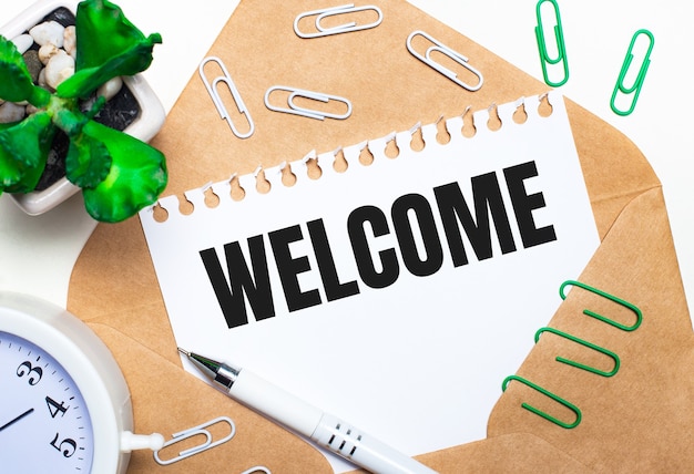 Photo on a light background, an open envelope, a white alarm clock, a green plant, white and green paper clips, a white pen and a sheet of paper with the text welcome