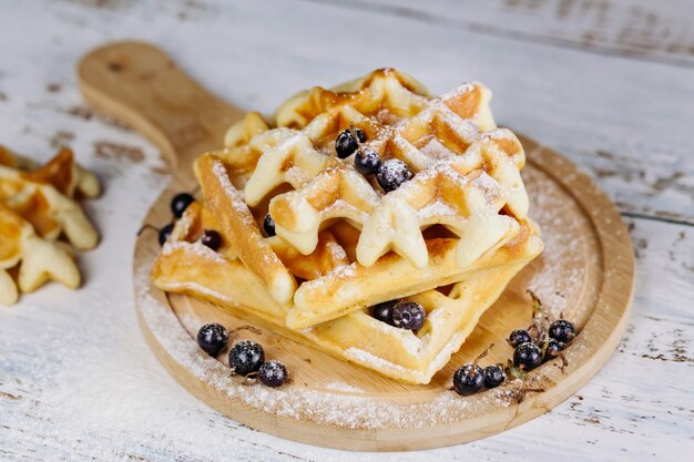 On a light background, Viennese waffles are stacked on a round board, one on one. Golden waffles are decorated with black currants.