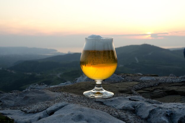 Light beer with froth in a glass stands on the edge of a hill against sunset sky close up. top view.