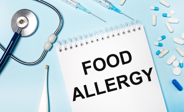 Su sfondo azzurro un termometro elettronico, uno stetoscopio, pillole bianche e blu, siringhe e un quaderno con la scritta food allergy. concetto medico