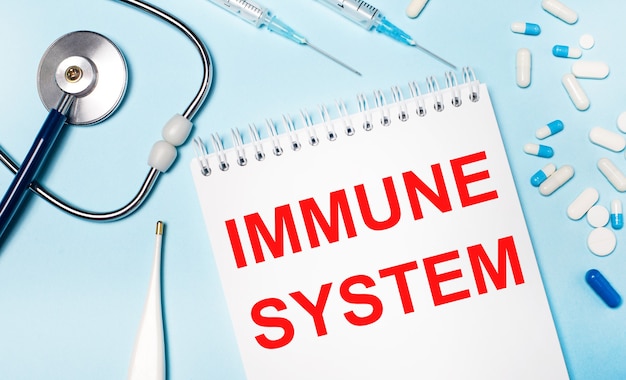 밝은 파란색 배경에 전자 온도계, 청진기, 흰색 및 파란색 알약, 주사기 및 IMMUNE SYSTEM이라는 텍스트가 있는 노트북. 의료 개념