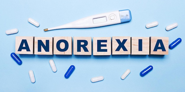 Su uno sfondo azzurro, un termometro elettronico, pillole bianche e blu e cubi di legno con la scritta anoressia. concetto medico