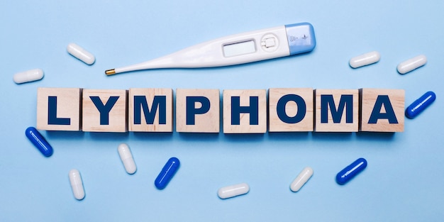 На голубом фоне электронный градусник, бело-синие таблетки и деревянные кубики с надписью LYMPHOMA. Медицинская концепция