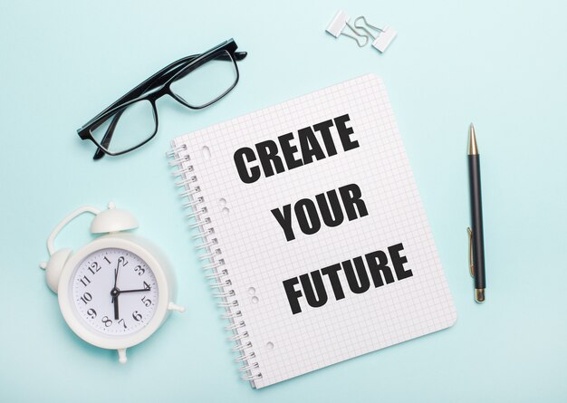 On a light blue background lie black glasses and a pen, a white alarm clock, white paper clips and a notebook with the words CREATE YOUR FUTURE