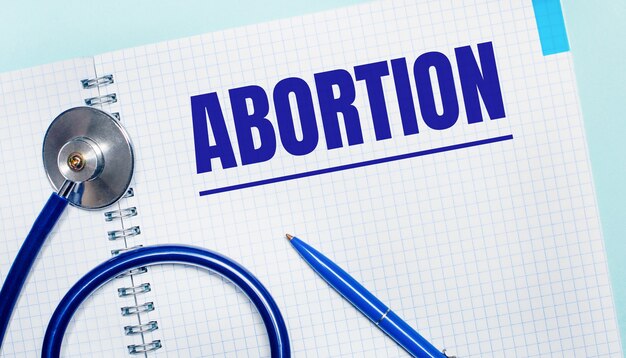 연한 파란색 배경에 ABORTION이라는 단어가 있는 노트북, 파란색 펜, 청진기가 있습니다. 위에서 볼. 의료 개념