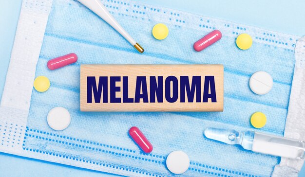 On a light blue disposable face mask there are tablets, a thermometer, an ampoule and a wooden block with the text MELANOMA. Medical concept