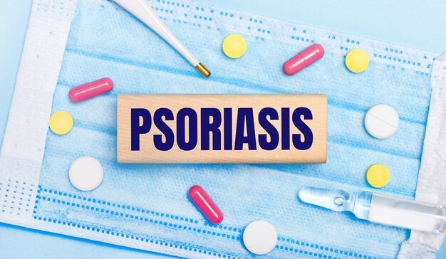 Su una mascherina usa e getta azzurra ci sono delle pastiglie, un termometro, una fiala e un blocco di legno con la scritta psoriasis. concetto medico