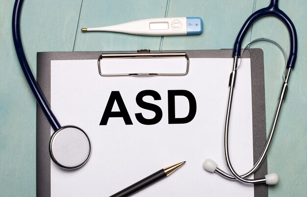 On a light blue wooden background, there is a paper labeled asd autism spectrum disorder, a stethoscope, an electronic thermometer, and a pen. medical concept