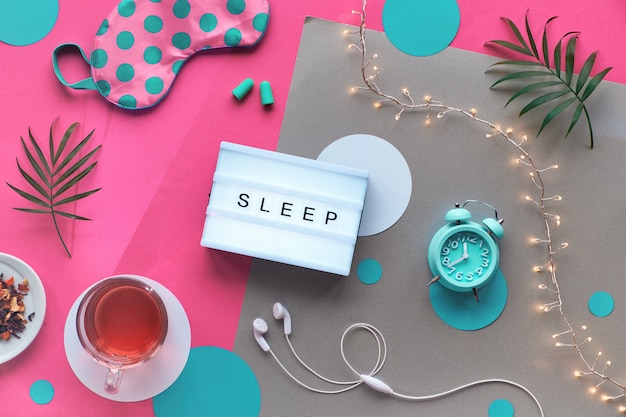 Light box, text "Sleep". Healthy night sleep creative flat lay. Sleeping mask, alarm clock, earphones, earplugs, herbal tea. Split pink and craft paper light garland and palm leaves