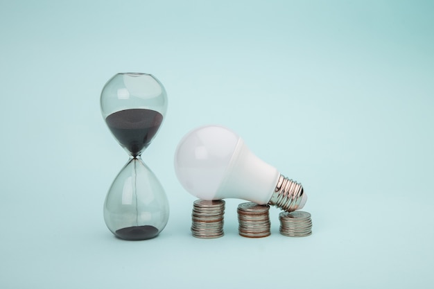 Light bulb with coins stack with hourglass on blue background. Power energy concept