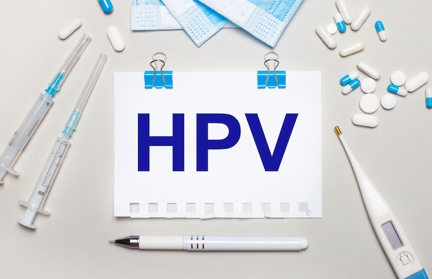 На светло-сером фоне синие медицинские маски, шприцы, электронный градусник, таблетки, ручка и блокнот с надписью HPV. Медицинская концепция