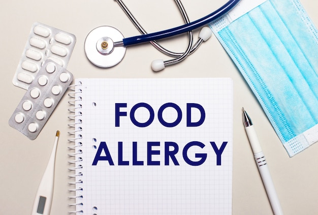 On a light gray background, a light blue disposable face mask, a stethoscope, an electronic thermometer, pills, a pen and a notebook with the inscription FOOD ALLERGY. Medical concept