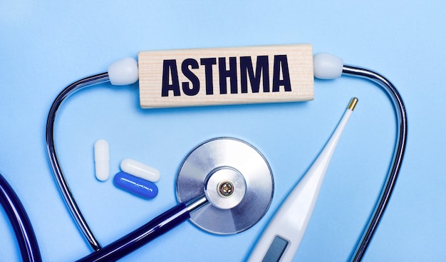 On a light gray background, a stethoscope, an electronic thermometer, pills, a wooden block with the text ASTHMA. Medical concept.