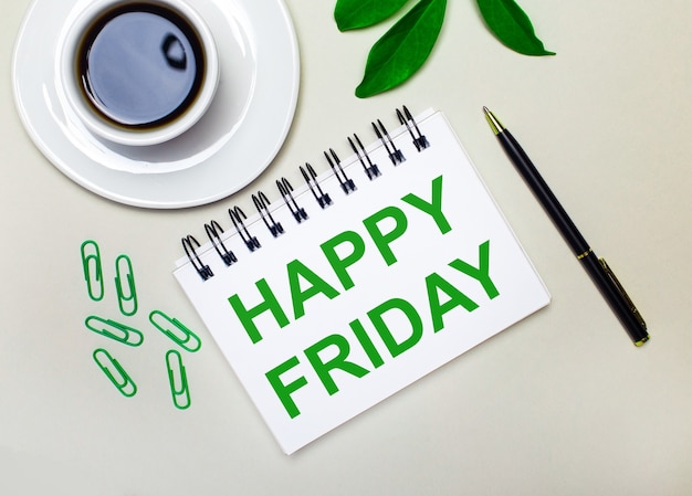 Photo on a light gray background, a white cup of coffee, green paper clips and a green leaf of a plant, as well as a pen and a notebook with the words happy friday.