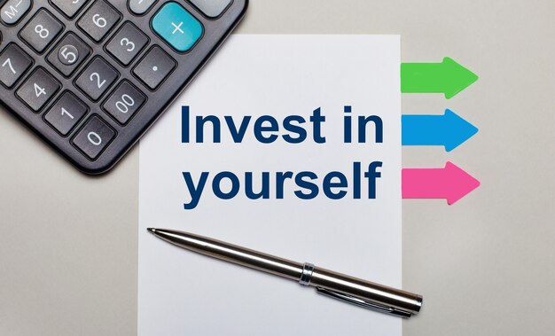 薄い灰色のテーブル、電卓、「INVEST IN YOURSELF」というテキストが書かれた白いシート、ペン、明るい色とりどりのステッカー。上から見る