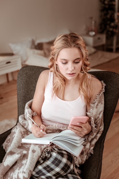 Light-haired beautiful woman. Busy fit lady sitting in armchair and listing down information from smartphone screen