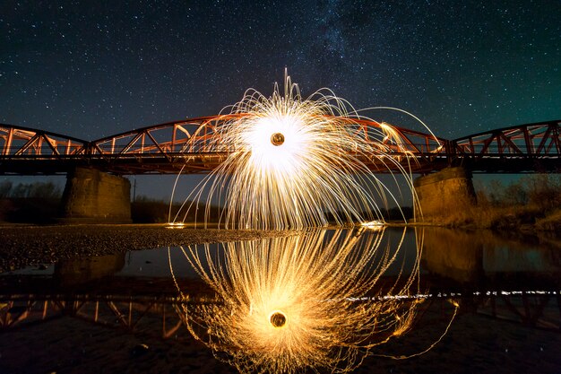 Light painting art concept. Spinning steel wool in abstract circle, firework showers of bright yellow glowing sparkles on long bridge reflected in river water on blue night starry sky