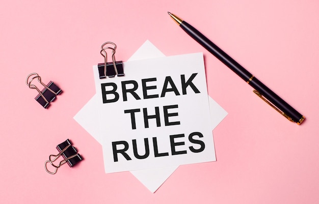 Photo on a light pink background, black paper clips, black pen and white note paper with the words break the rules. flat lay