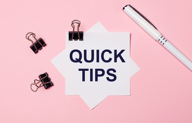 Photo on a light pink background, black paper clips, black pen and white note paper with the words quick tips. flat lay