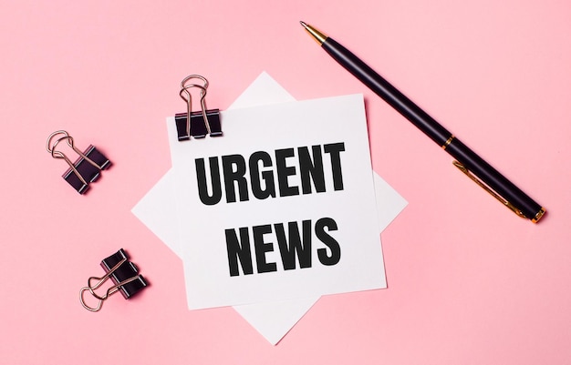 On a light pink background, black paper clips, black pen and white note paper with the words URGENT NEWS. Flat lay