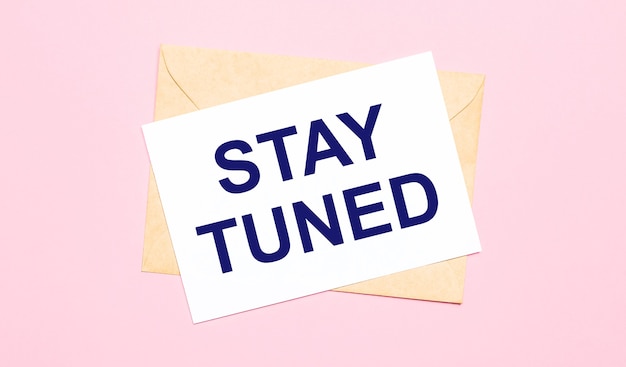 On a light pink background - a craft envelope. It has a white sheet of paper that says STAY TUNED