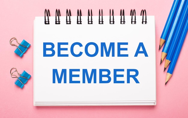 밝은 분홍색 배경에 밝은 파란색 연필, 종이 클립 및 BECOME A MEMBER라는 텍스트가 있는 흰색 공책