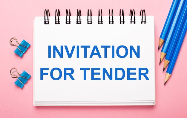 On a light pink background light blue pencils paper clips and a white notebook with the text INVITATION FOR TENDER