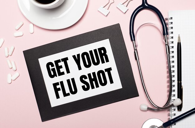 On a light pink background, a notebook with a pen, stethoscope, white pills, paper clips and a sheet of paper with the text GET YOUR FLU SHOT. Medical concept