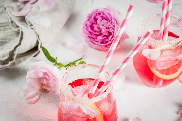 Rosa chiaro rosa cocktail, con vino rosato, petali di rosa tea e limone su un tavolo di cemento bianco pietra.