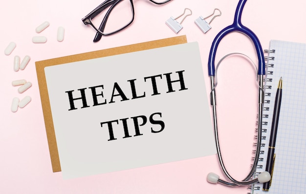 On a light pink surface, a stethoscope, white pills and clips for paper, glasses in black frames and a sheet of paper with the text HEALTH TIPS