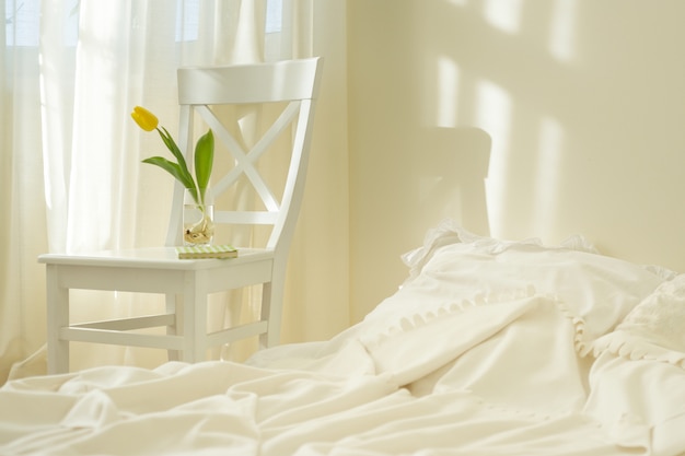 Light spring bedroom interior, bed, white chair, glass with yellow tulip