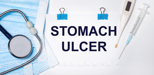 On a light surface lie a stethoscope, an electronic thermometer, a syringe, a face mask and a sheet of paper with the text STOMACH ULCER