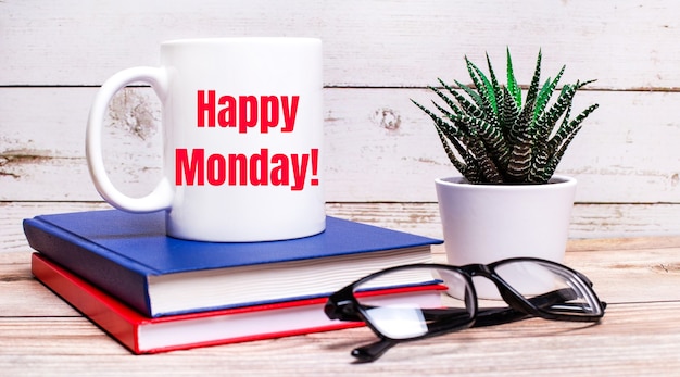 Photo on a light table are notepads, a potted plant, black-framed glasses and a white cup with the text happy monday.