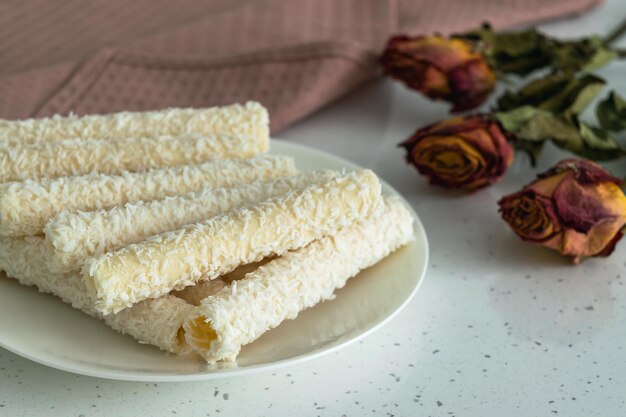 On a light table, sweets for tea and coffee on a porcelain plate. Wafer rolls with coconut flakes and cream next to a bouquet of dried flowers and a powdered napkin.