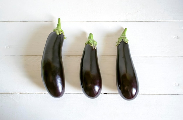 On a light table there are three ripe eggplants Three eggplants lie in a row on a white wooden table