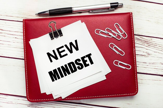 밝은 나무 배경에 버건디 펜과 노트북 노트북에는 NEW MINDSET Business concept이라는 텍스트가 있는 흰색 종이 클립과 흰색 종이가 있습니다.