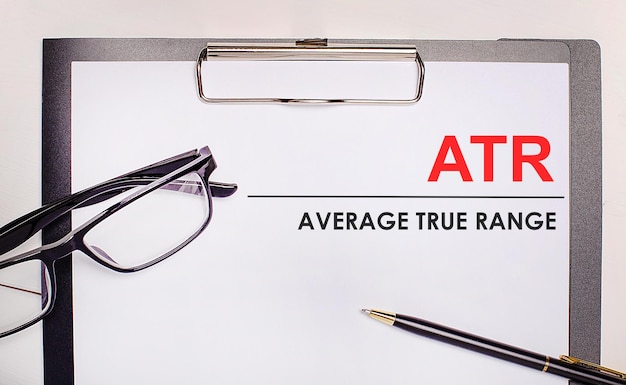 On a light wooden background glasses a pen and a sheet of paper with the text ATR Average True Range Business concept