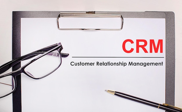 On a light wooden background glasses a pen and a sheet of paper with the text CRM Customer Relationship Management Business concept