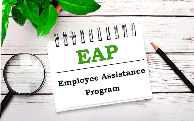 On a light wooden background a magnifying glass a pencil a green plant and a white notebook with text EAP Employee Assistance Program Business concept