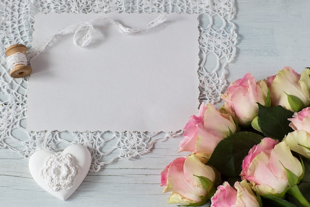 on a light wooden background a sheet of paper, pink roses, a heart, rose petals 